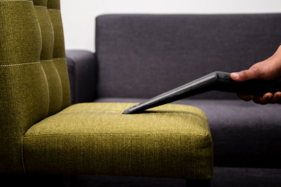 A close-up of a hand using a vacuum cleaner attachment to thoroughly clean the mustard-yellow upholstered seat of a modern sofa, with a dark grey fabric sofa in the background. The scene is well-lit, 