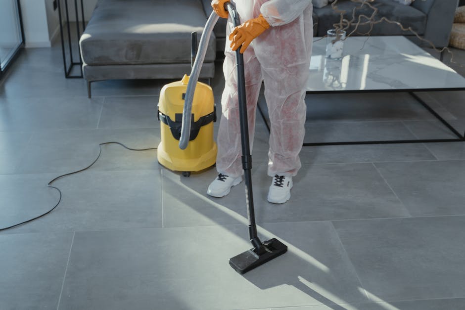 A close-up of a hand using a vacuum cleaner attachment to thoroughly clean the mustard-yellow upholstered seat of a modern sofa, with a dark grey fabric sofa in the background. The scene is well-lit, 