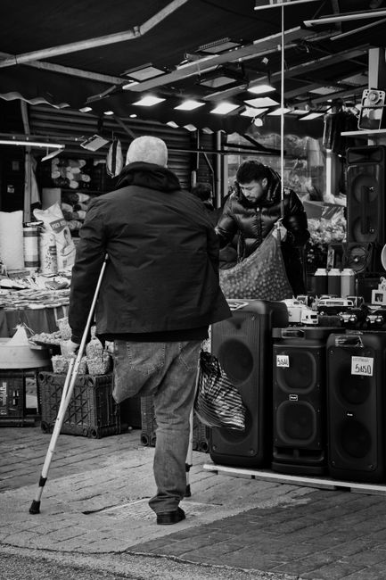 The image depicts an indoor market setting with a variety of tables and displays, featuring items for sale. The scene includes several market traders, with one prominent man in the foreground wearing 